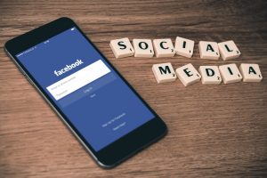 Auf einem Smartphone ist die Facebook-App geöffnet. Neben dem Smartphone ist mit Scrabble Steinen die Wörter "Social Media" geschrieben.