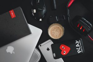 Laptop mit Tasche, Notebook, Uhr, Kopfhörer, Smartphone mit Hülle und eine Tasse Kaffee vor schwarzem Hintergrund.