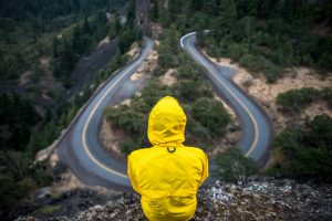 Eine PErson mit einer gelben Kapuzenjacke sitzt auf einem Berg und schaut herab auf eine gewundene Straße